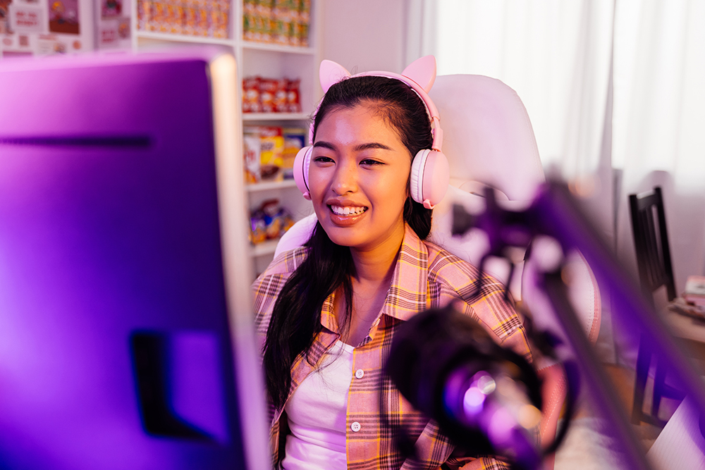young woman wearing a pink cat eat headset smiles while looking at her computer screen with a microphone in the foreground